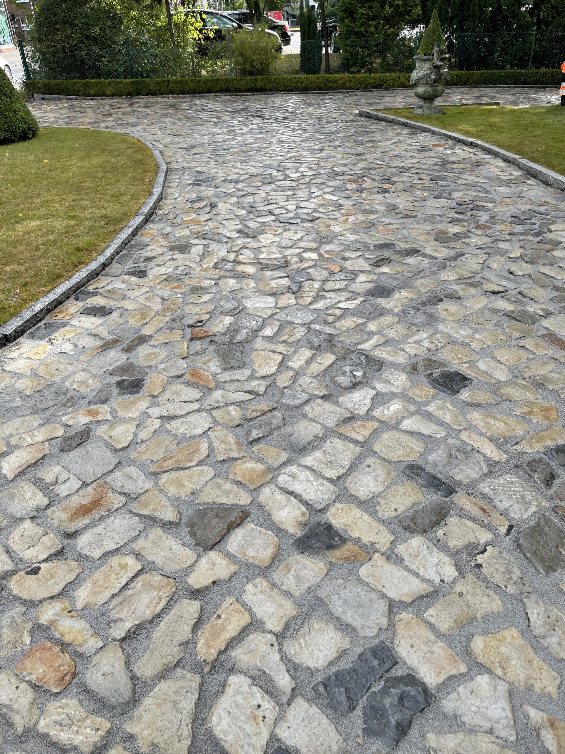 Gepflasterter Weg aus unregelmäßigen hellgrauen und beigen Steinen führt kurvig zwischen gepflegten Rasenflächen, begrenzt durch niedrige Granitkanten und Hecken mit Pflanzen im Hintergrund.