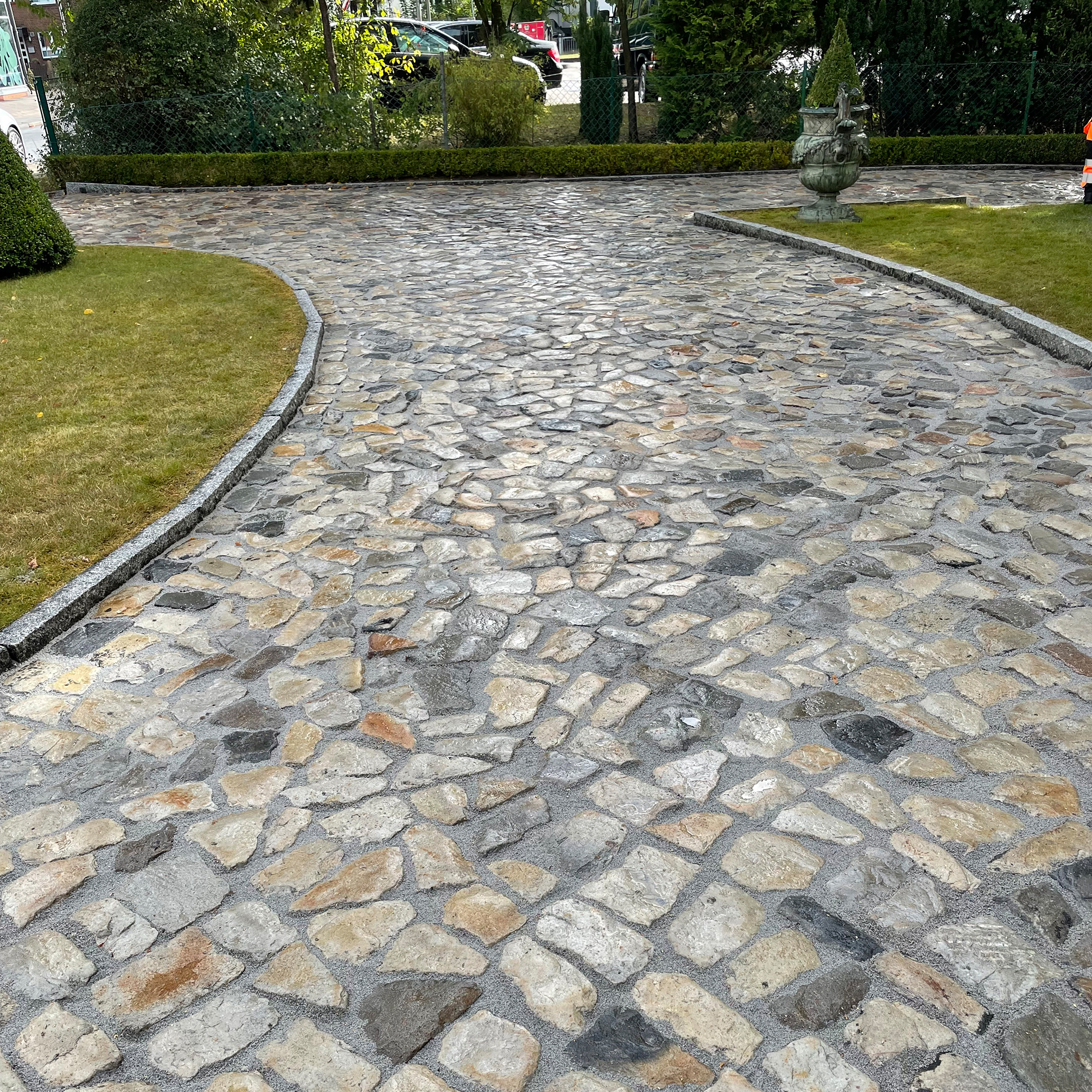 Kopfsteinpflasterweg, leicht nass, verläuft in sanfter Kurve; beidseitig von niedrigen Granitkanten eingefasst, angrenzende Rasenflächen, Hecke und große Zierurne im Hintergrund.