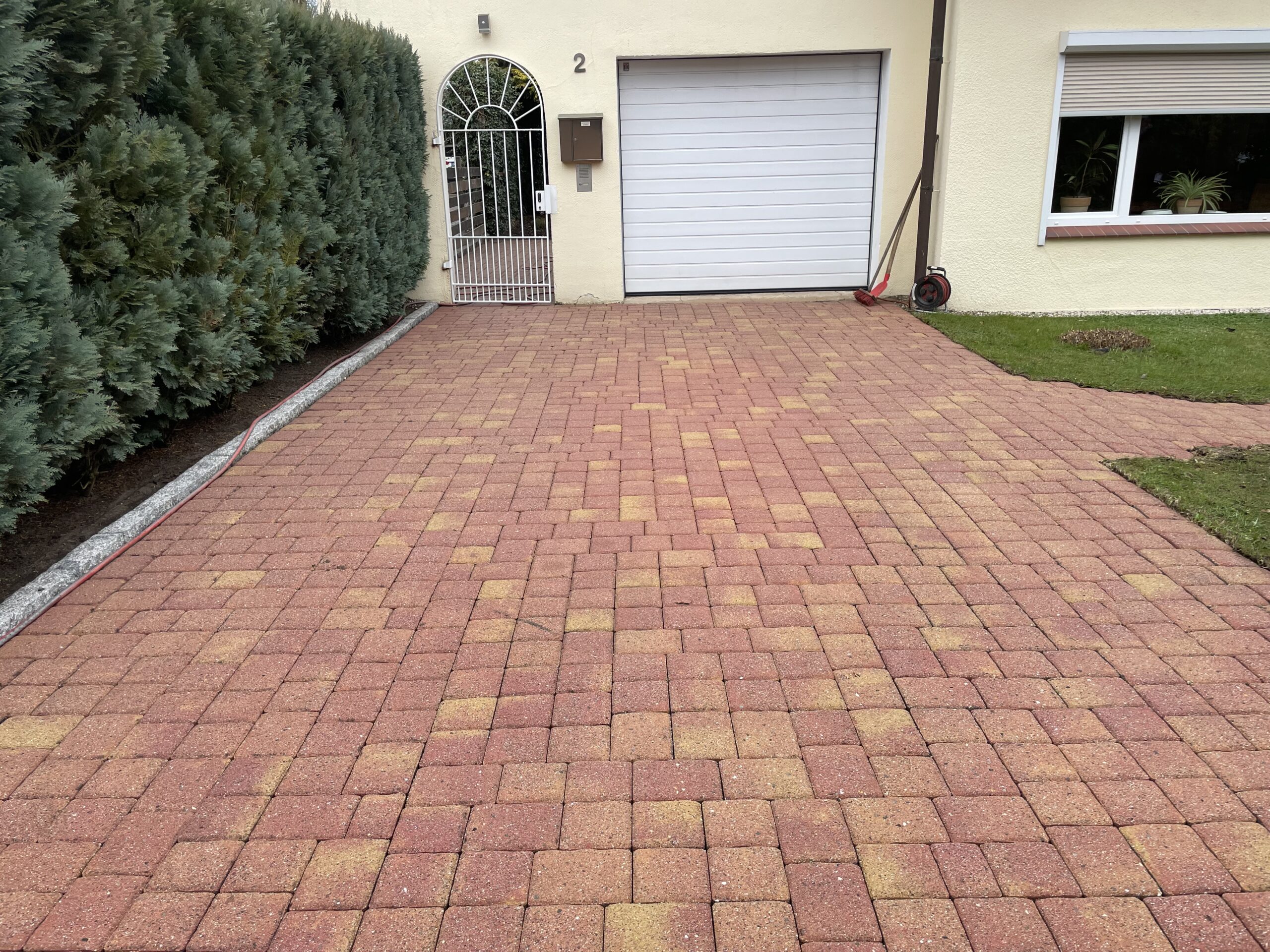 Rote gepflasterte Einfahrt, die zu geschlossener weißer Garagentür und weißem Eingangstor mit Hausnummer 2 führt; links dichte Hecke, rechts Rasen, Fenster mit Pflanzen, Schlauchrolle.