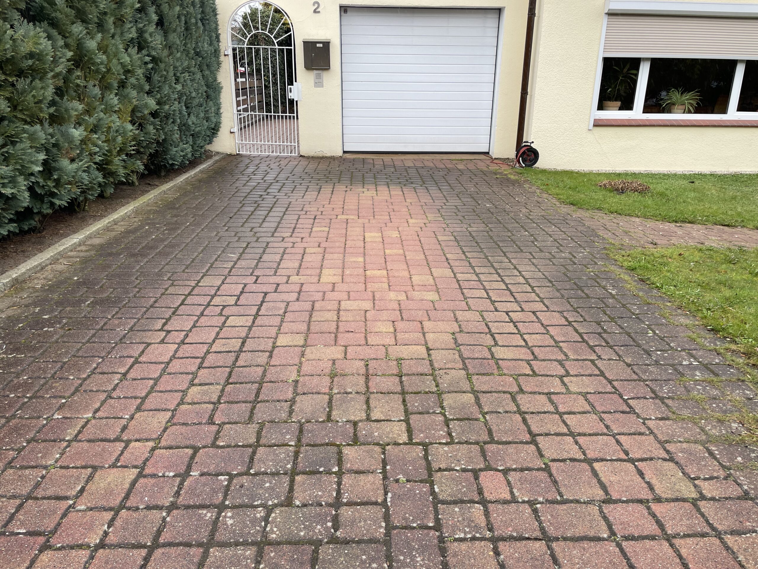 Gepflasterte Einfahrt – verläuft zur geschlossenen weißen Garagentür und einem weißen Tor mit Hausnummer "2" – flankiert von dichter Hecke links, Rasen und Fenster mit Pflanzen rechts.
