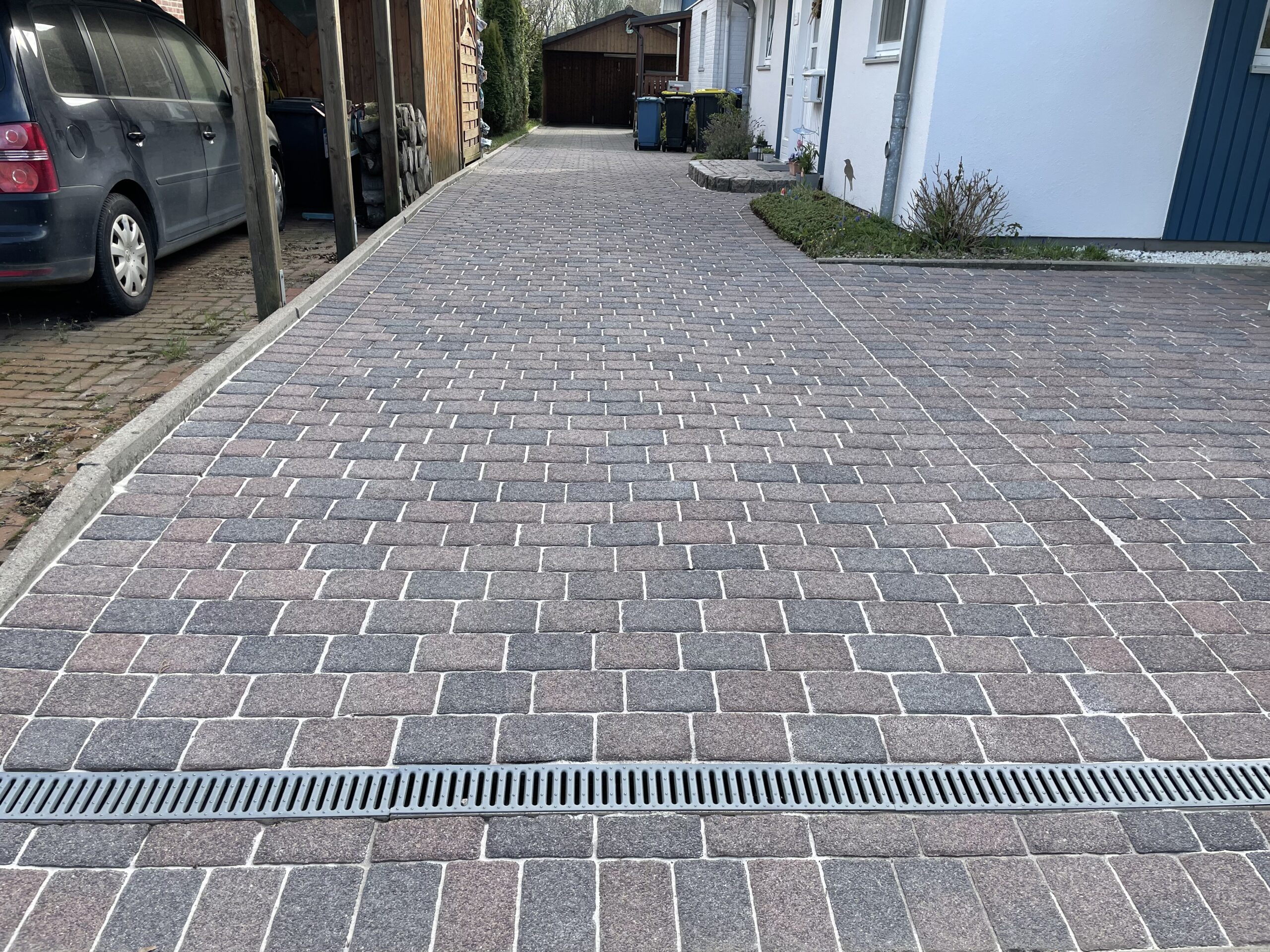 Pflasterauffahrt mit rechteckigen grauen und roten Klinkerpflastersteinen, führt zur Garage; Metall-Rinnenrost quer im Vordergrund, links Carport mit geparktem dunklem Auto, rechts Hausfassade und Mülltonnen.