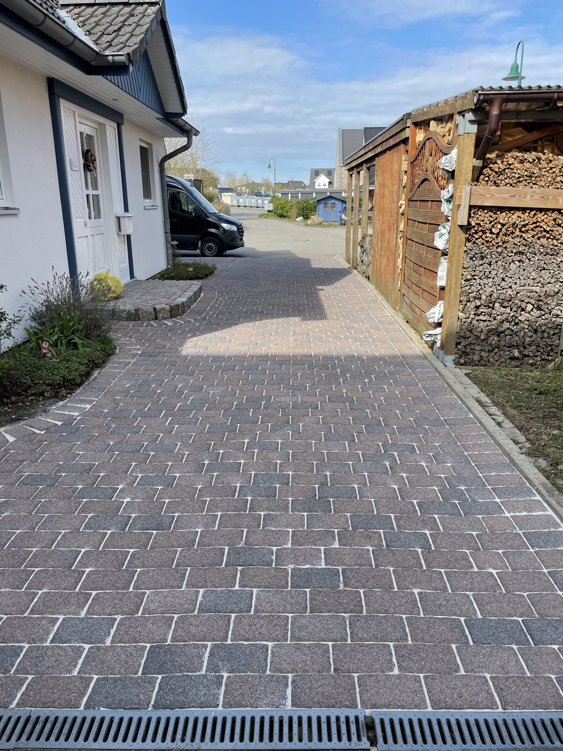 Gepflasterte Einfahrt, die zur Straße führt; links weißes Haus mit Haustür und schwarzem Transporter geparkt, rechts Holzschuppen mit gestapeltem Brennholz, blauer Himmel.
