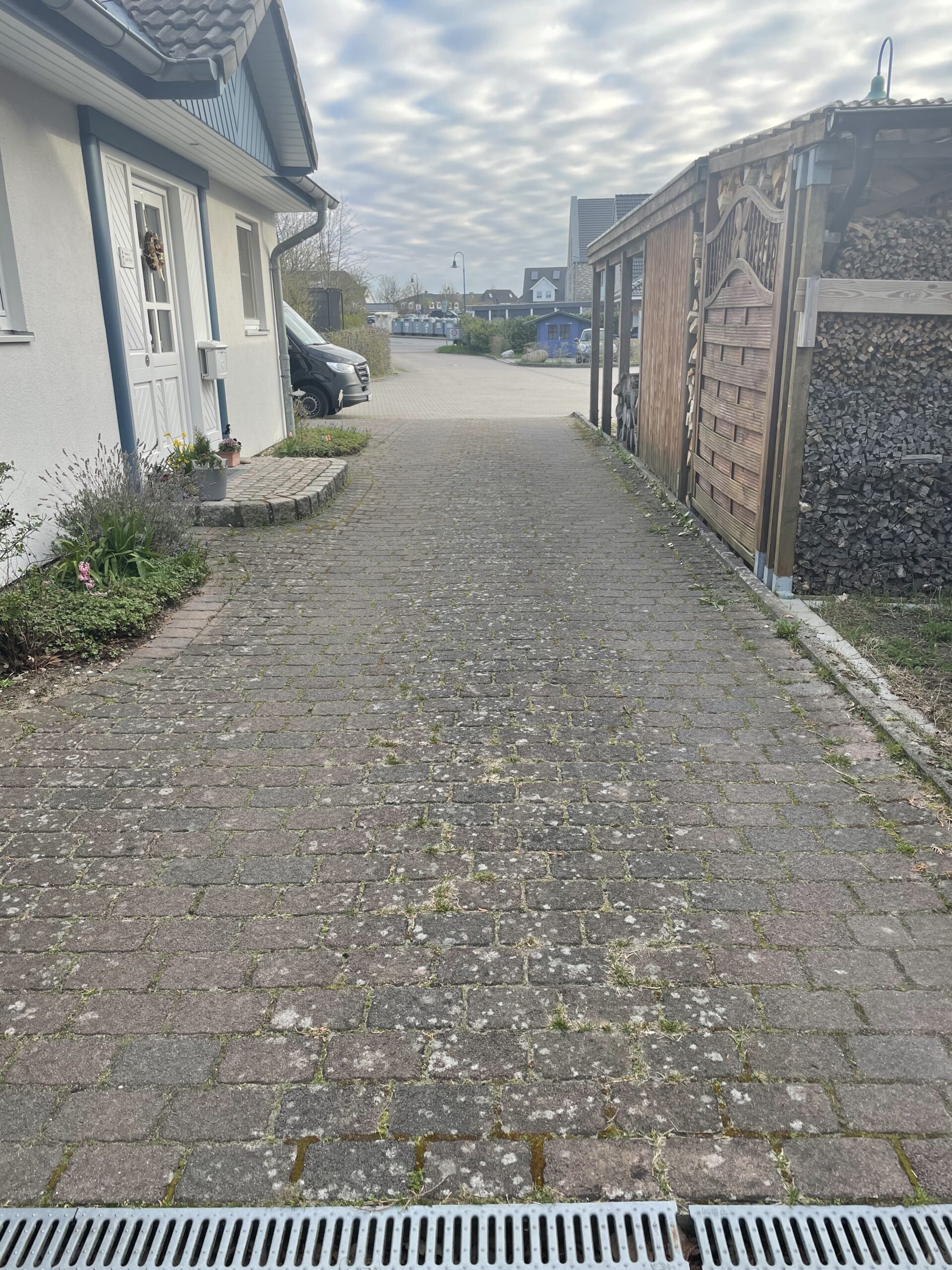 Gepflasterte Auffahrt mit Entwässerungsrinne im Vordergrund verläuft zur Straße, zwischen einem weißen Haus mit Haustür und Blumentöpfen links und einem Holzschuppen mit gestapeltem Brennholz rechts, Lieferwagen am Ende.