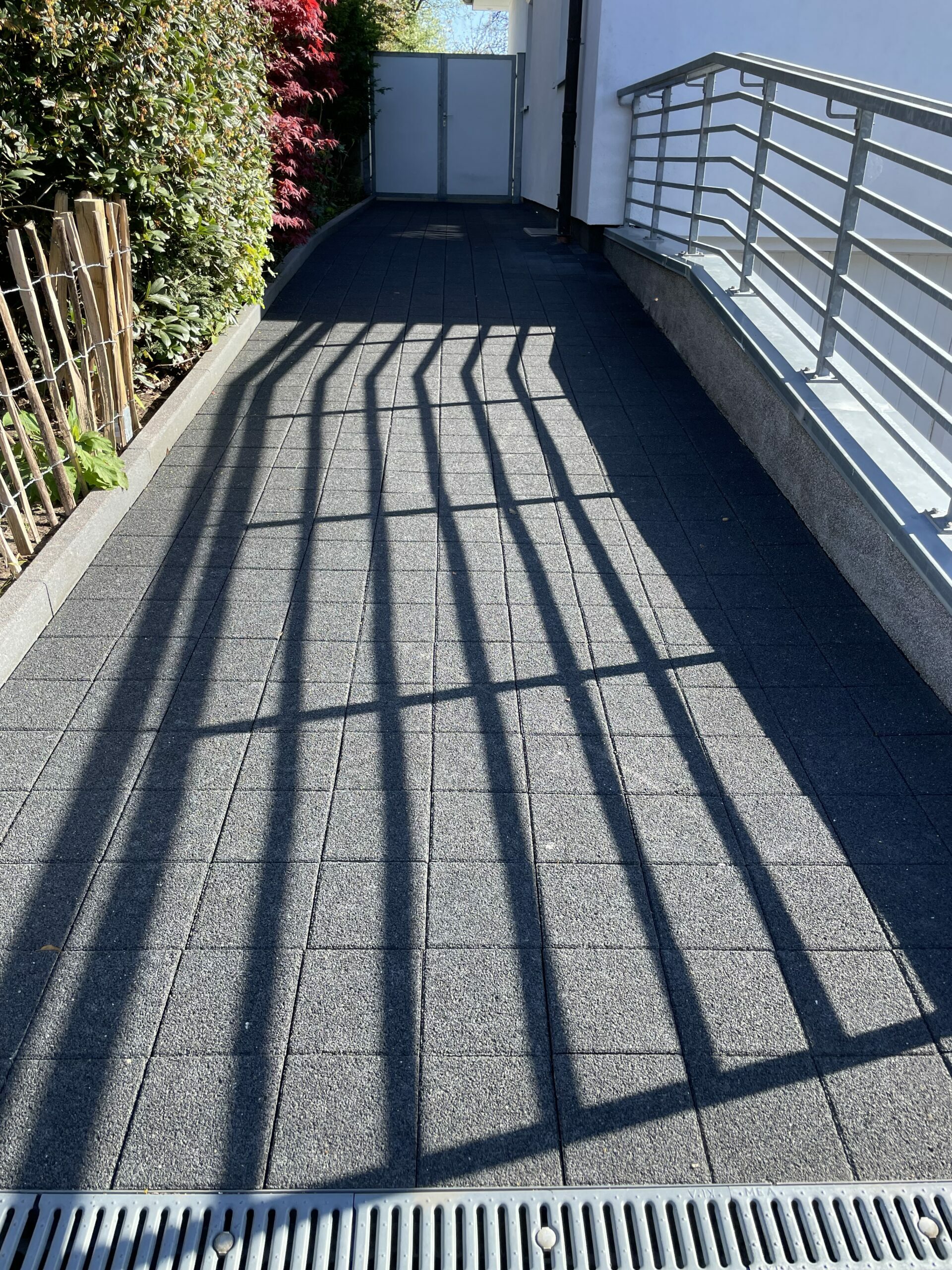 Metallgeländer wirft lange, parallele Gitter‑Schatten auf dunkelgraue quadratische Pflastersteine; links dichte Hecke und Bambuszaun, rechts weiße Hausfassade mit Handlauf und geschlossenem Tor.