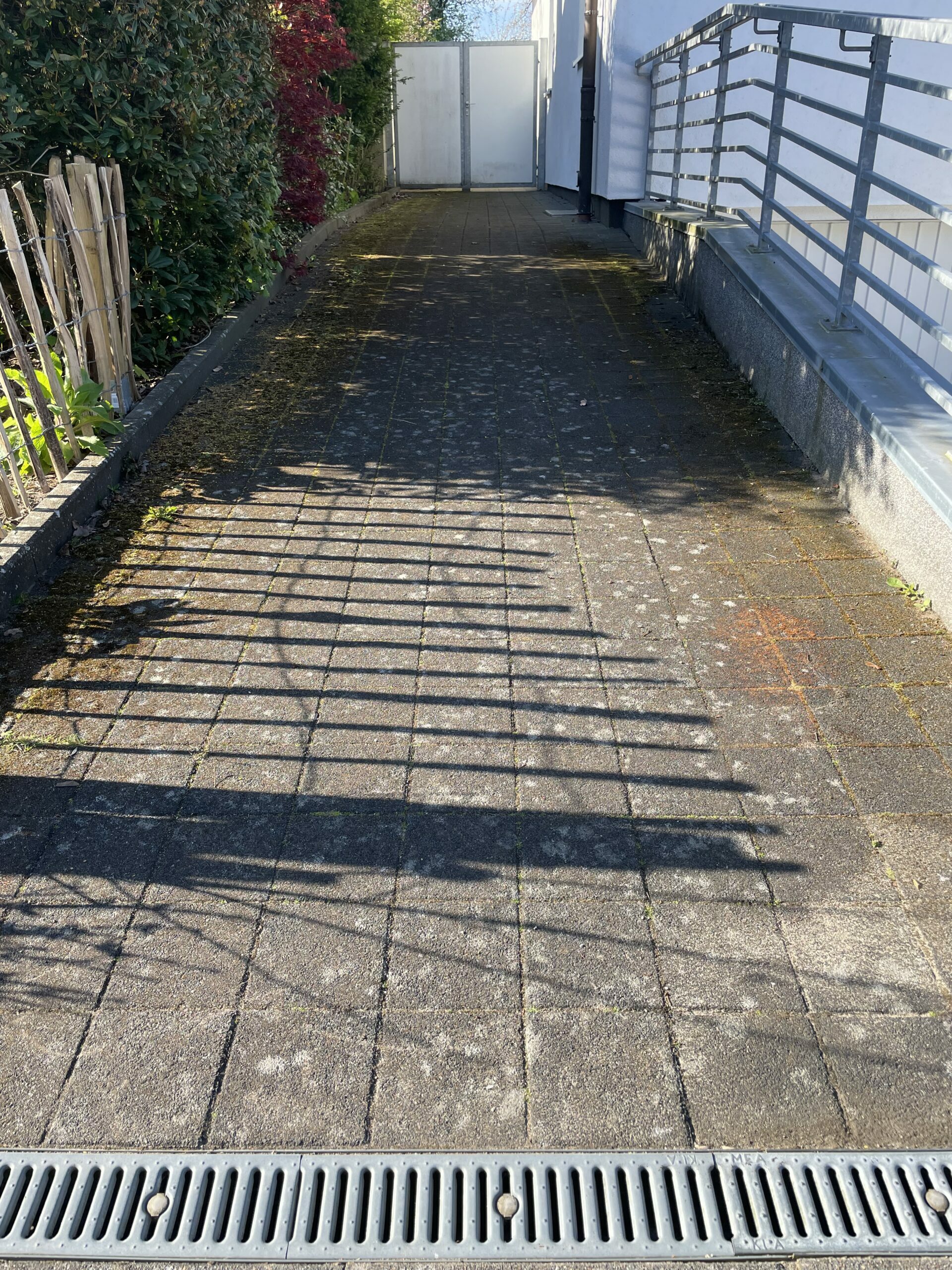 Pflasterweg – Schatten eines Metallgeländers fallen als parallele Streifen auf die Platten; links dichte Hecke mit Holzlatten, rechts Hauswand mit Geländer, weißes Tor am Ende.