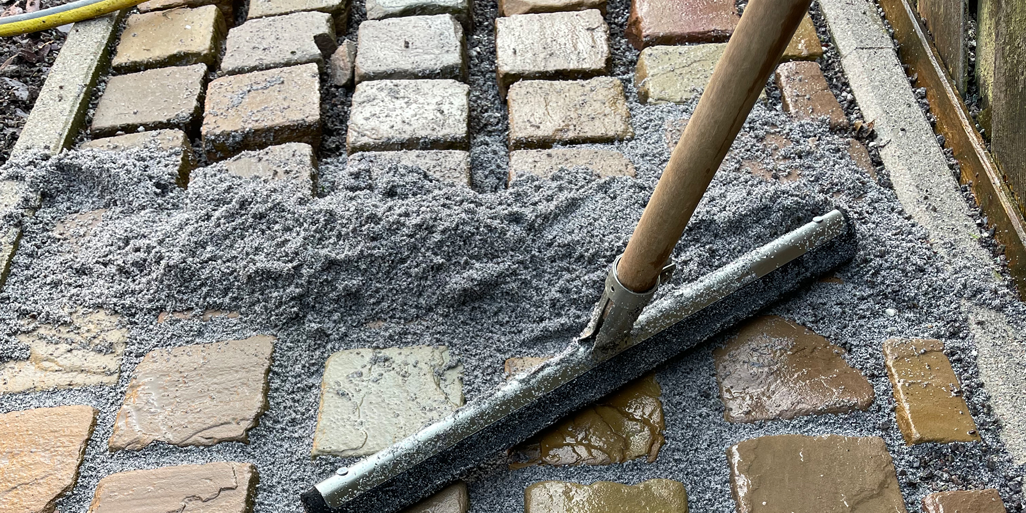 Schrubber mit Holzstiel und schwarzer Gummilippe schiebt graues, feinkörniges Material über nasse, quadratische Pflastersteine, verteilt in Fugen nahe Rand und Holzzaun.