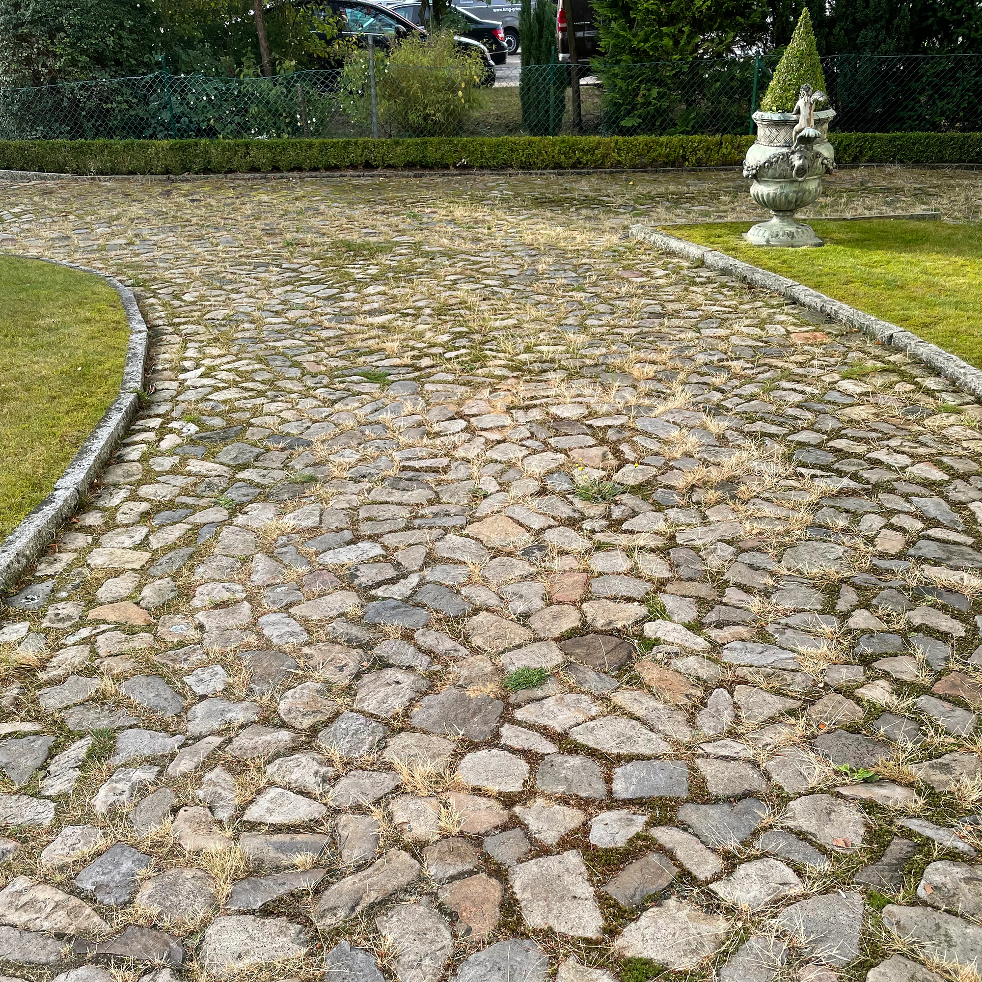 Pflasterweg aus unregelmäßigen Kopfsteinpflastersteinen verläuft geschwungen, trockenes Gras wächst zwischen den Fugen, gesäumt von Rasen, rechts verzierte Steinurne, hinten Hecke und parkende Autos.