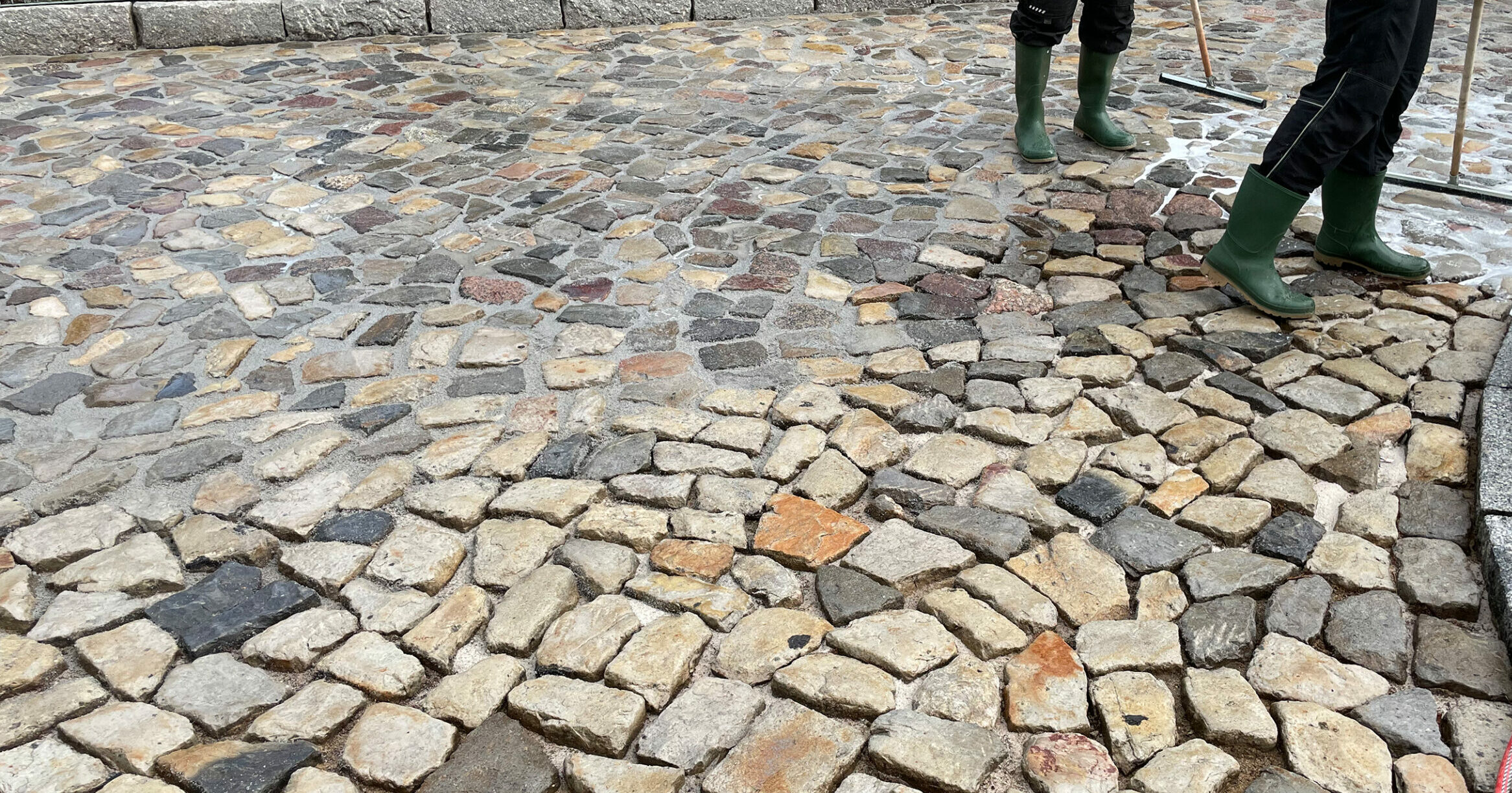 Unregelmäßiges Kopfsteinpflaster wird mit Wasser und Gummirakeln gereinigt; nasser mehrfarbiger Steinbelag, zwei Personen mit grünen Gummistiefeln und schwarzer Arbeitskleidung am Rand.