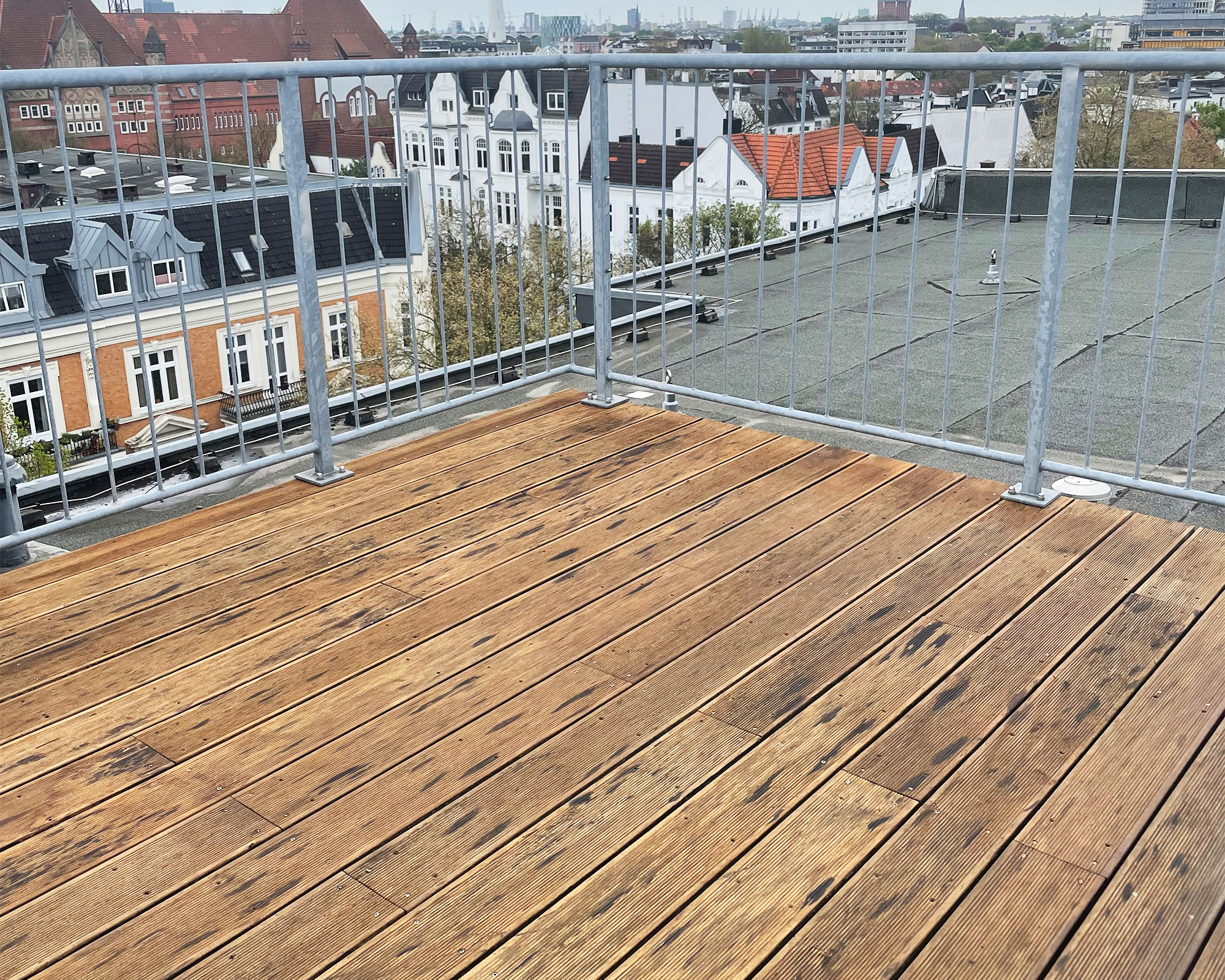 Holzterrassendielen erstrecken sich im Vordergrund bis zu einem Geländer aus Metall, das auf die Dächer und Fassaden mehrerer Stadthäuser unter einem bewölkten Himmel blickt.
