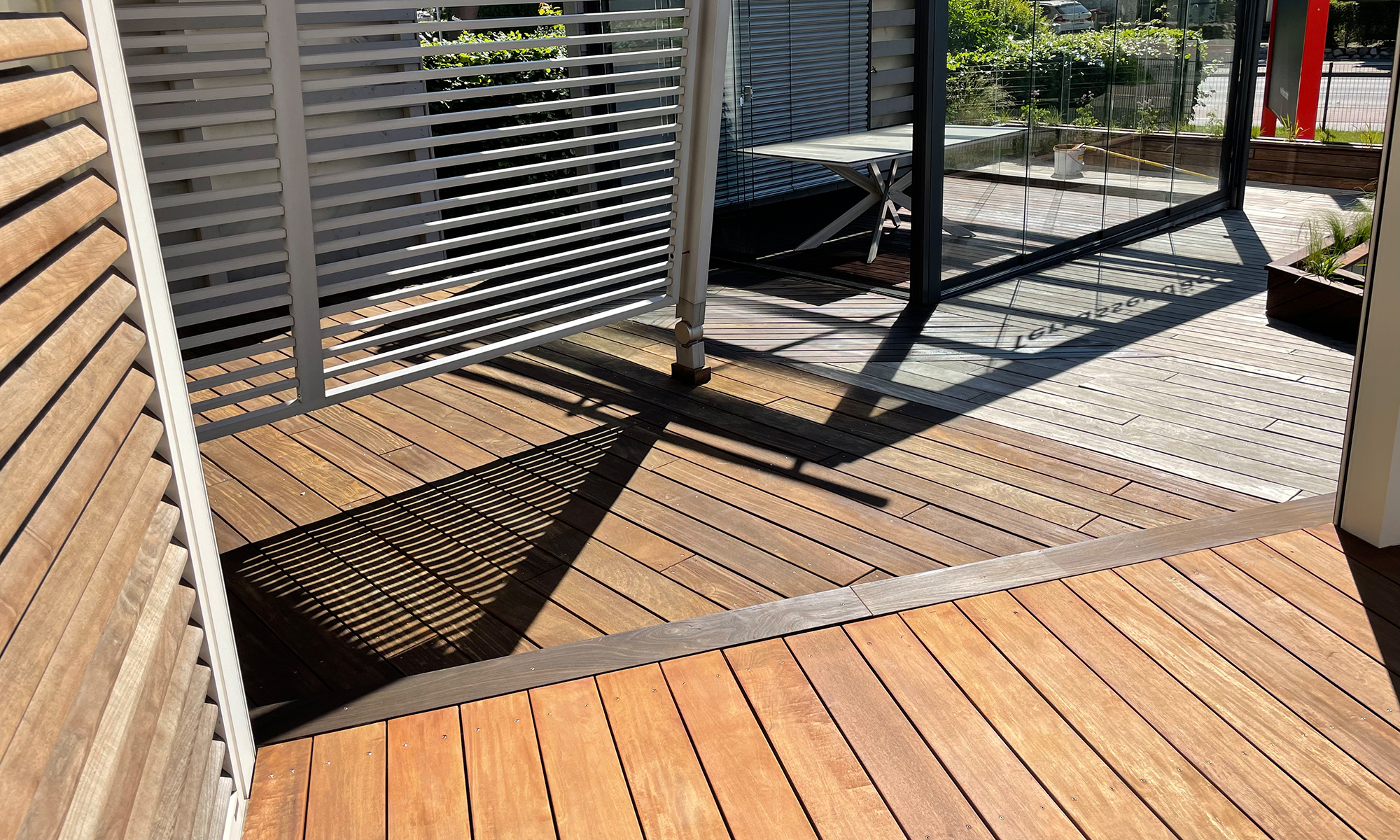 Weiße horizontale Lamellenwand wirft ein schattiertes Streifenmuster auf hellbraune Holzdielen; sonnige, teilweise verglaste Terrasse mit klappbarem Tisch und Pflanzkästen im Hintergrund.
