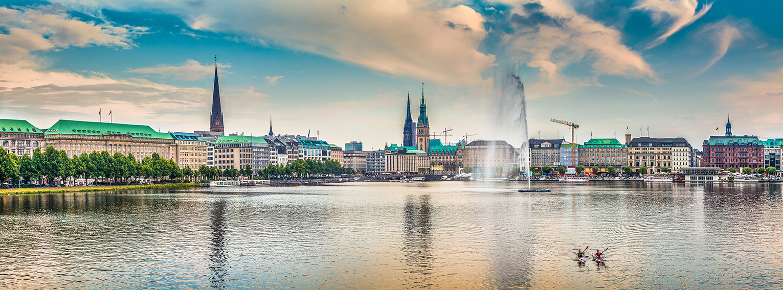 Stadt-Silhouette am See mit hoher Fontäne, die Wasser in die Luft spritzt; zwei Kajakfahrende paddeln im Vordergrund, Ufer mit Bäumen, historischen Gebäuden und Kirchtürmen.