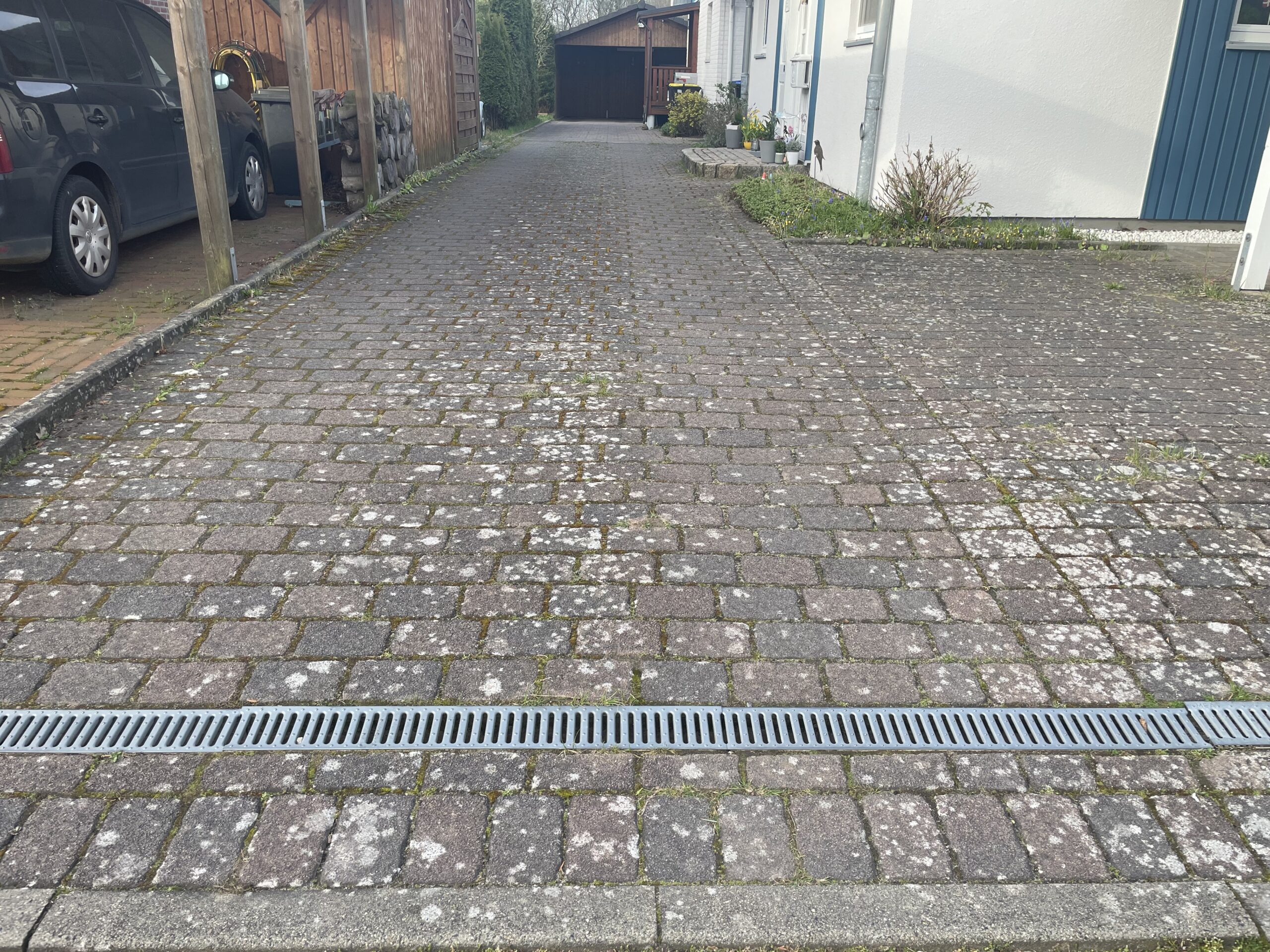 Pflasterauffahrt mit Metall-Drainagerinne im Vordergrund, bedeckt von Moos- und Flechtenflecken; links Carport mit geparktem Auto, rechts Hauswand, Vorgarten und Garage im Hintergrund.